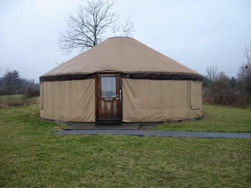 A tan yurt with a wooden door, set in a grassy area with sparse trees and a cloudy sky in the background.