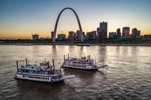 Two steamboats on a river with a city skyline and arch monument in the background during sunset.