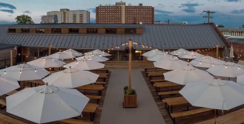 Outdoor dining area with wooden tables, white umbrellas, and string lights, set against a city skyline at dusk.