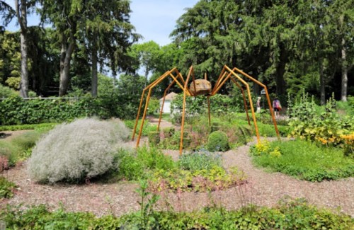 A large metal spider sculpture in a lush garden surrounded by greenery and colorful plants.