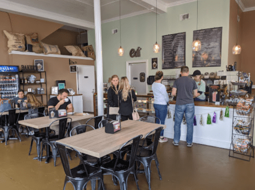 A cozy café interior with patrons at tables and a counter, featuring a menu board and decorative elements.