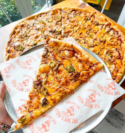 A hand holding a large slice of pizza with jalapeños, next to a whole pizza on a wooden table.