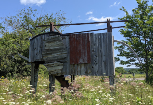 A rustic wooden structure resembling a bull, set in a grassy field with trees and blue sky in the background.