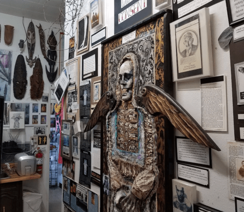 A wall filled with various artworks and framed images, featuring a central sculpture of a winged figure.