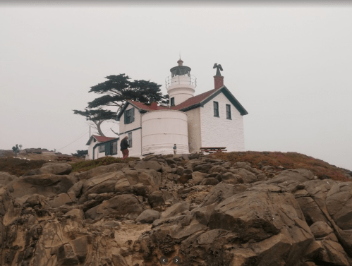 A white lighthouse with a red roof stands on rocky terrain, surrounded by fog and greenery.
