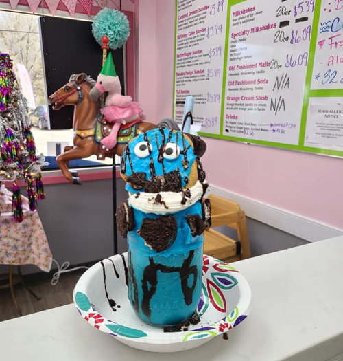 A colorful dessert in a glass, topped with whipped cream, cookies, and chocolate drizzle, with a playful horse decoration in the background.