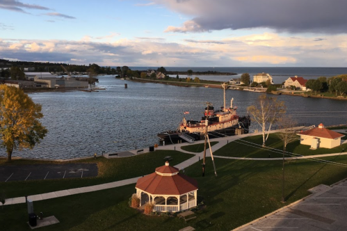 A scenic view of a river with a tugboat, gazebo, and trees under a partly cloudy sky.