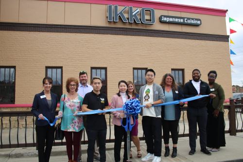 A group of people gathers outside a restaurant, holding a blue ribbon for a grand opening ceremony.