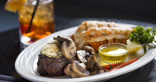 A gourmet plate featuring filet mignon with mushrooms, lobster tail, lemon, and a side of dipping sauce.