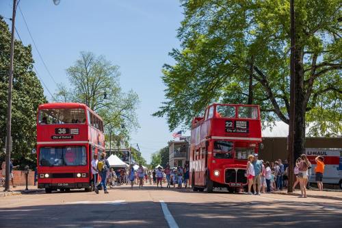 Two red double-decker buses on a sunny street, with people walking and trees lining the road.