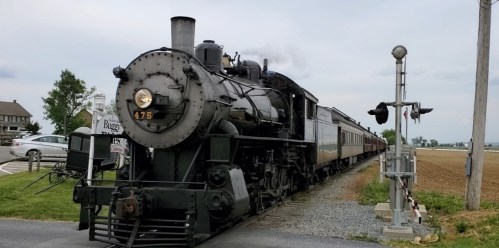 A vintage steam locomotive with passenger cars stops near a railroad crossing in a rural setting.