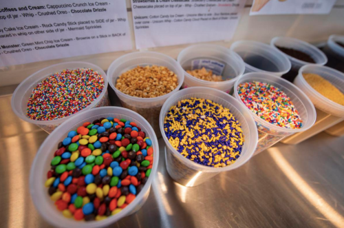 Colorful sprinkles and toppings in clear cups, including chocolate chips, nuts, and rainbow sprinkles.