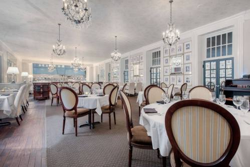 Elegant restaurant interior with chandeliers, round tables set with white linens, and framed artwork on the walls.