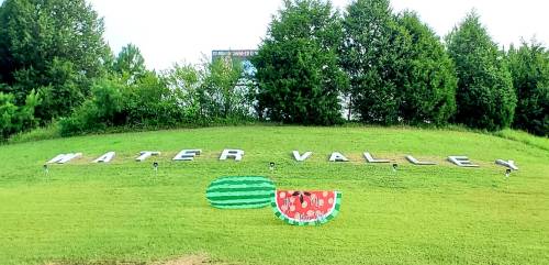 Sign reading "WATER VALLEY" on a grassy hill, with colorful watermelon decorations on either side.
