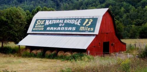 Red barn with a large sign reading "Natural Bridge of Arkansas 13 Miles" surrounded by greenery and hay bales.