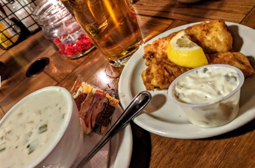 A plate with fried fish and a lemon wedge, accompanied by a cup of clam chowder and a small container of tartar sauce.