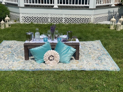A cozy outdoor setup with a low table, drinks, and decorative pillows on a patterned rug in a grassy area.