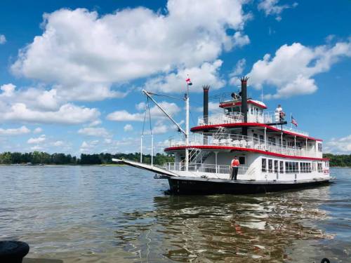 A large riverboat with red and white accents docked on a calm river under a blue sky with fluffy clouds.