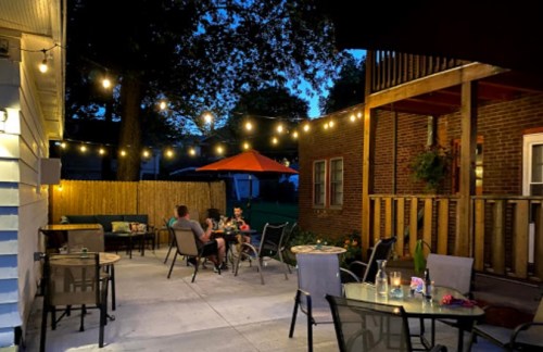 A cozy outdoor patio at night, with string lights, tables, and a couple enjoying a meal under an orange umbrella.
