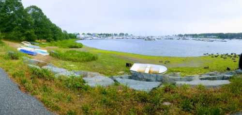 A serene waterfront scene with boats on the shore, grassy areas, and a calm bay in the background under a cloudy sky.