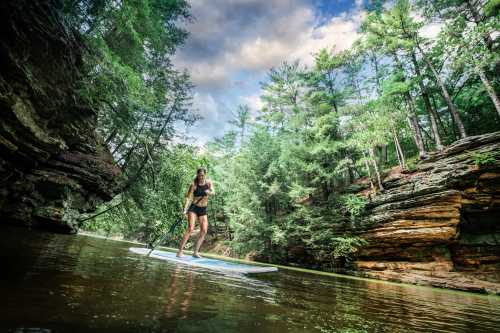 A woman paddleboards on a calm river surrounded by lush green trees and rocky cliffs under a cloudy sky.