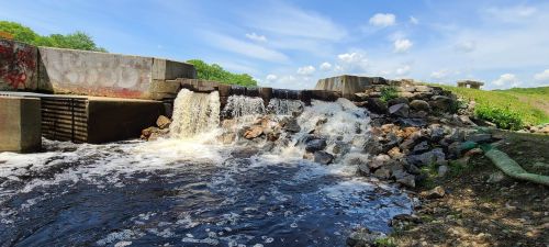 A small dam with water cascading over rocks, surrounded by greenery and a blue sky with scattered clouds.