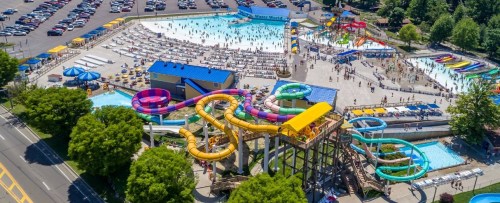 Aerial view of a busy water park with colorful slides, pools, and sunbathers, surrounded by trees and parking.
