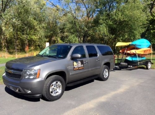 A gray SUV parked beside a trailer carrying colorful kayaks in a natural outdoor setting.