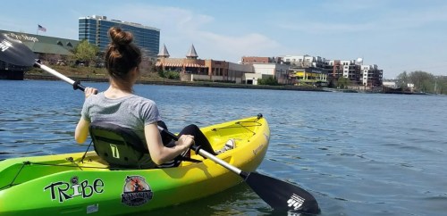 A person in a kayak on a calm river, with a city skyline and buildings in the background under a clear blue sky.