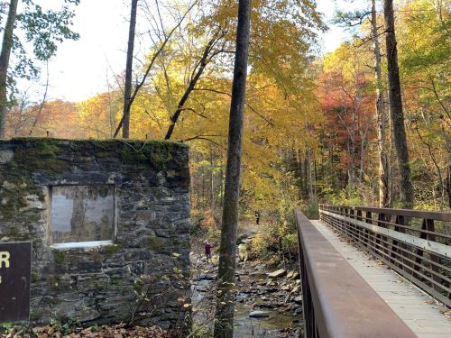 A stone structure beside a creek, with a wooden bridge and vibrant autumn foliage in the background.