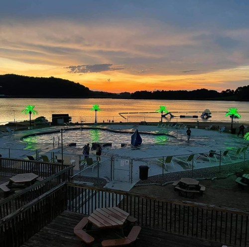 Sunset over a pool with green palm tree lights, surrounded by water and mountains, with people enjoying the scene.