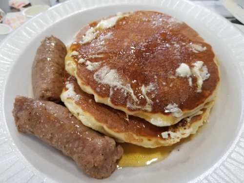 A plate with three fluffy pancakes topped with butter and syrup, alongside two sausage links.