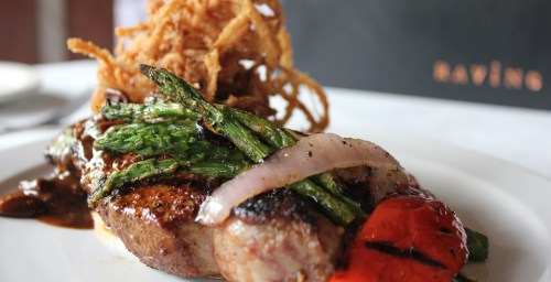 A plated steak topped with grilled asparagus, onions, and roasted red peppers, garnished with crispy onion strings.