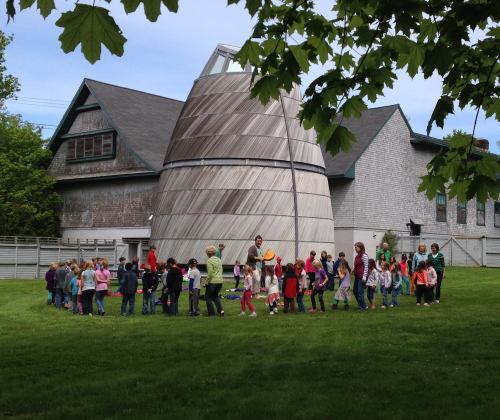 A group of children and adults gather around a unique, modern building on a grassy area, with trees in the foreground.