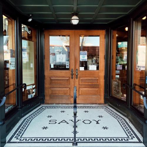 Entrance of a building with wooden double doors and a tiled floor that reads "SAVOY." Signs indicate it's open.