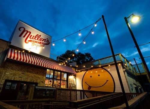Mullen's Dairy Bar at dusk, featuring a smiling mural and string lights illuminating the outdoor seating area.