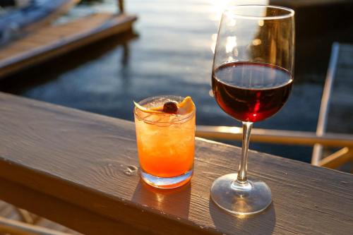 A glass of red wine and a cocktail with orange and cherry sit on a wooden railing by the water at sunset.