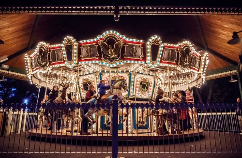 A brightly lit carousel with ornate horses, surrounded by a fence, at night with people enjoying the ride.