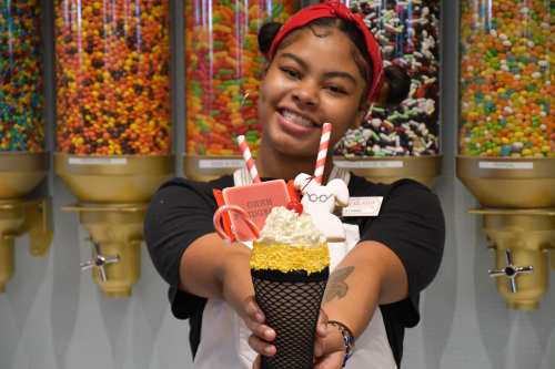 A smiling young woman holds a colorful ice cream treat in front of a candy-filled display.