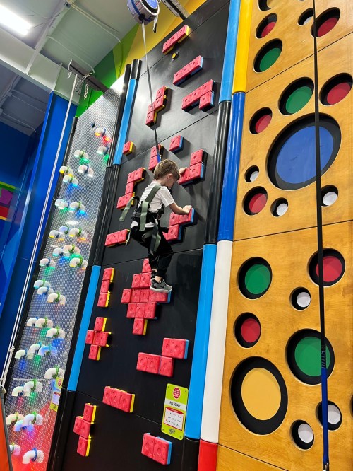 A child climbs a colorful indoor rock wall with various shapes and holds, wearing a safety harness.
