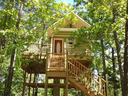 A charming treehouse nestled among green trees, featuring a red door and a wooden staircase leading up to it.