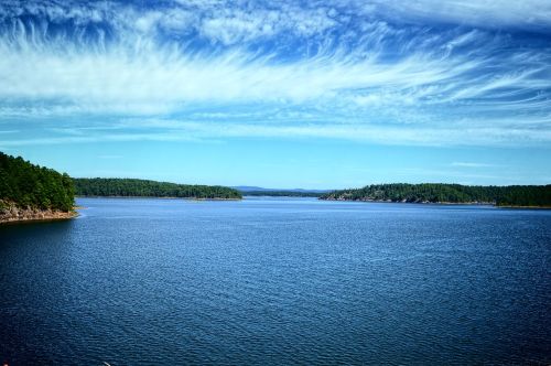 A serene lake surrounded by lush green hills under a blue sky with wispy clouds.