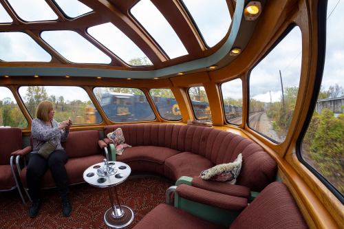 A woman sits in a cozy train lounge with large windows, as another train passes by outside.