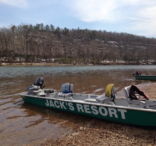A green boat with "JACK'S RESORT" on the side is docked by a river, surrounded by trees and rocky terrain.