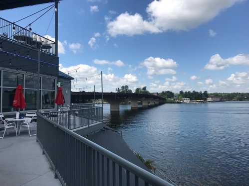 A view of a river with a bridge in the background, featuring a patio with red umbrellas and blue sky with clouds.