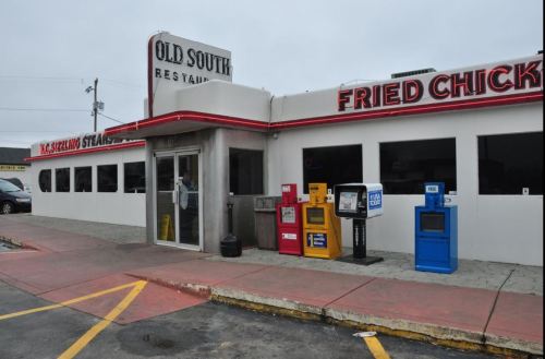 Exterior of Old South Restaurant featuring a sign for fried chicken and newspaper boxes in front. Cloudy sky above.