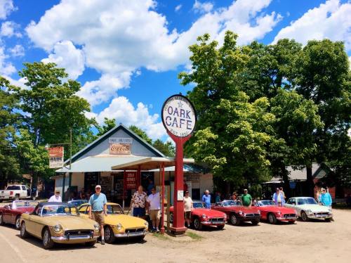 A vintage café with a sign, surrounded by classic cars and people enjoying a sunny day.