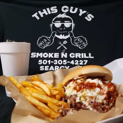 A delicious BBQ sandwich with coleslaw and fries, alongside a drink, on a table with a restaurant logo in the background.