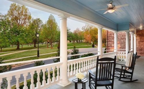 A porch with rocking chairs overlooks a lush green park with trees and a pathway.