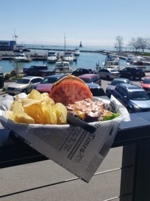 A burger with tomato and lettuce served with chips, overlooking a marina filled with boats and parked cars.